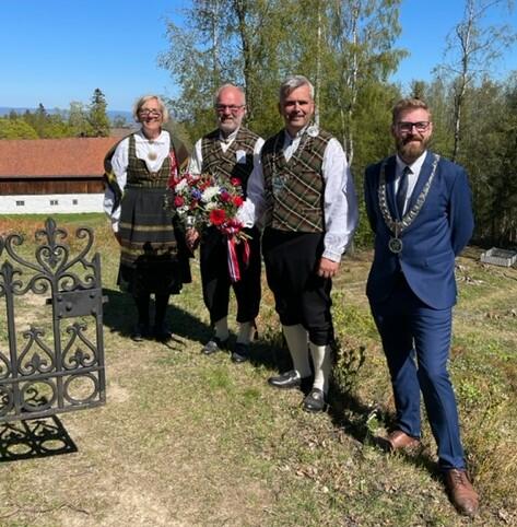 Fire personer, festpyntede representanter for Toten historielag og ordførere i Østre og Vestre Toten står ved gravstedet til Weidemannfamilien, med en blomsterkrans som skal legges på graven. Sol og blå himmel. Låven på Stenberg i bakgrunnen.