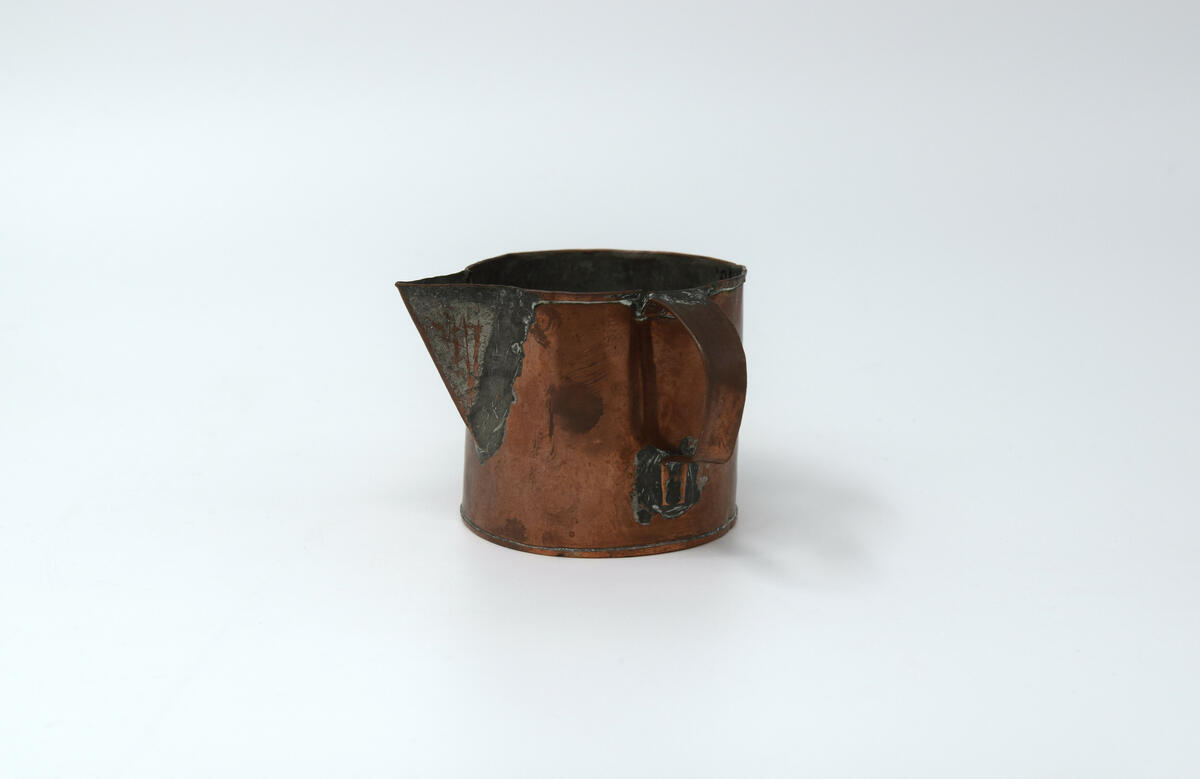 Kannans botten är rund och har rakt sidostycke med en smal fals i överkant och en pip 2,5 cm djup. Pipen är tennlödd och förtent. Handtaget är utfört i vikt plåt och fästad med tennlödning. Insidan är förtent. Botten är plan o med tennlödd fals sidostycket har slaglödd fals till höger om handtaget. Tillstånd: Överkanten är bucklig. En reva på var sida om handtaget.