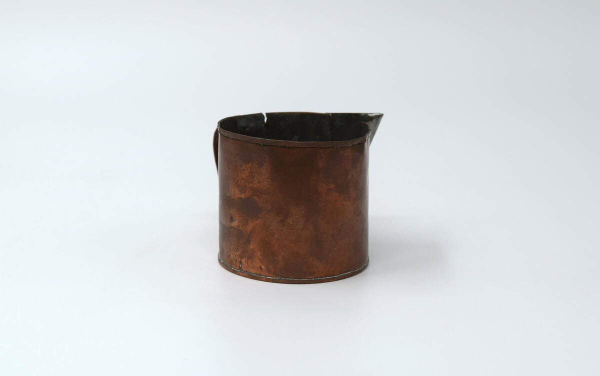 Kannans botten är rund och har rakt sidostycke med en smal fals i överkant och en pip 2,5 cm djup. Pipen är tennlödd och förtent. Handtaget är utfört i vikt plåt och fästad med tennlödning. Insidan är förtent. Botten är plan o med tennlödd fals sidostycket har slaglödd fals till höger om handtaget. Tillstånd: Överkanten är bucklig. En reva på var sida om handtaget.