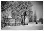 En snöig väg med en putsad tvåvåningsbyggnad bakom en mur och höga träd.