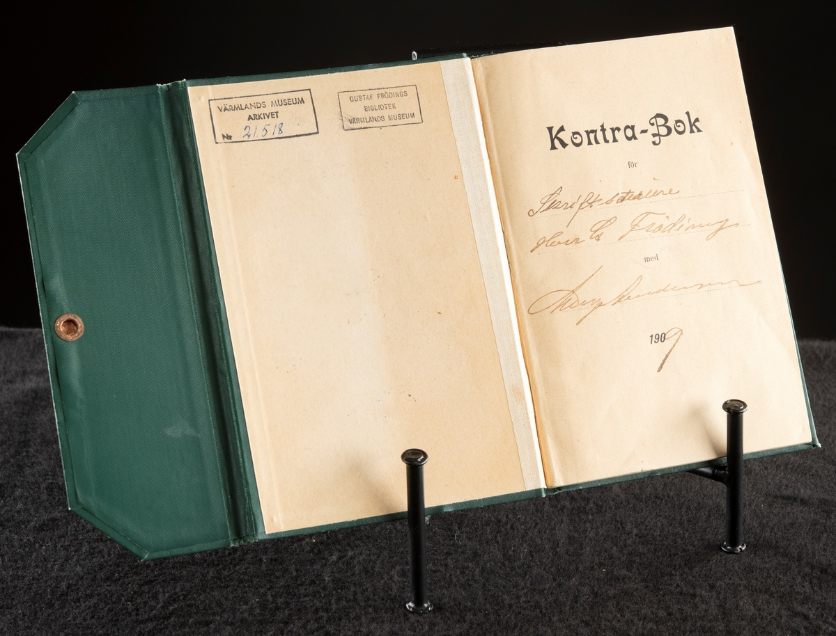 Grönt klot, med extra omslag för att stänga boken med tryckknapp på baksidan.
Präglad pärm med blommönstrad ram. Texten i guldprägling.