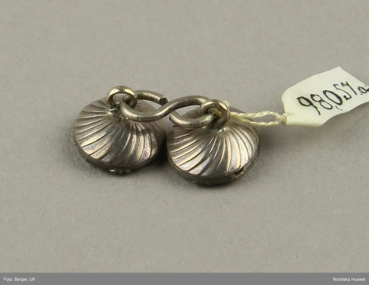 Manschettknappar, två par, miniatyrer. 1700-tals mitt.  Silver, glas.
Saknar stämplar. På varje knapp ett ovalt porträtt i miniatyr i silverinfattning, baksidan räfflad, knapparna förenade två och två, på den ena föreningslänken. ”I H” i genombrutit arbete, den andra föreningslänken senare ersättning. Manschettknapparna hölls i ett rött marokäng (getskinn) etui.
(katalogiseringen gäller a-c)