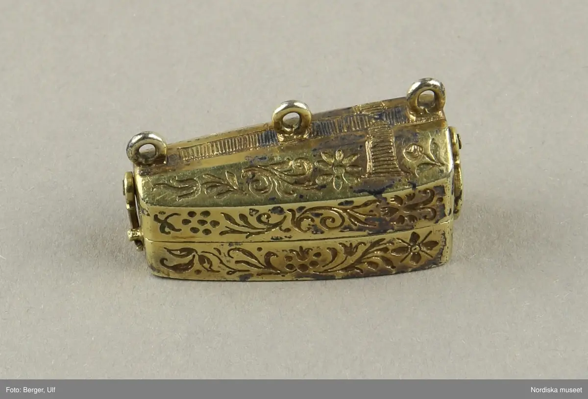Katalogkort:
"Smycke (berlock)
Silver
L 3,5 cm, br. 1,2 cm
I form av Gustav II Adolfs likkista. Trycklock med haspar. Graverad dekor. Miniatyrbild i trä av kunge, fäst i botten."