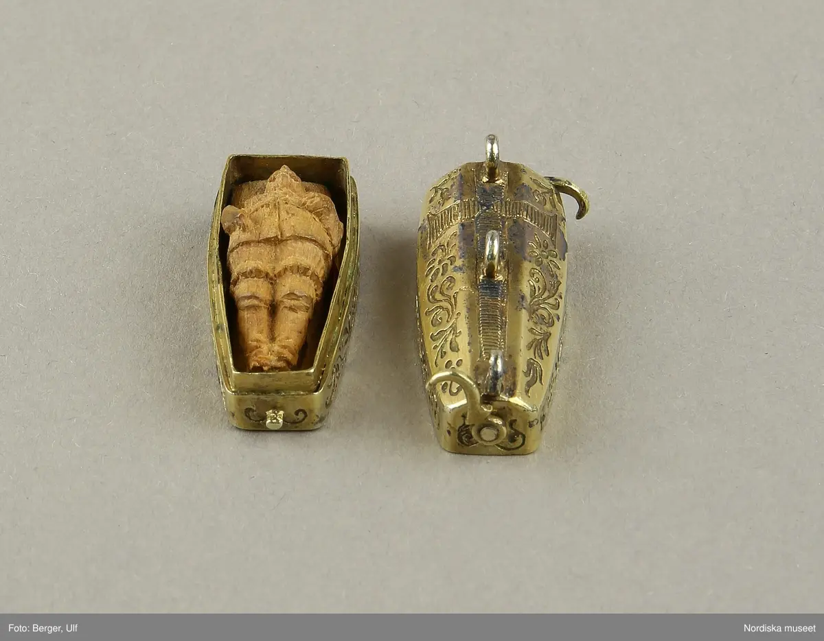 Katalogkort:
"Smycke (berlock)
Silver
L 3,5 cm, br. 1,2 cm
I form av Gustav II Adolfs likkista. Trycklock med haspar. Graverad dekor. Miniatyrbild i trä av kunge, fäst i botten."