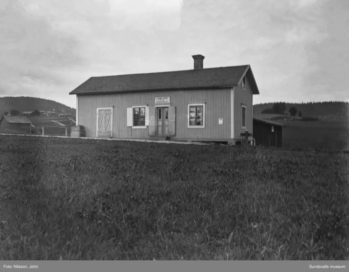 En ljus, låg byggnad med skylt över entrédörren: I A Hallbergs Diversehandel. Till vänster i bakgrunden syns en lantgård med flera byggnader.