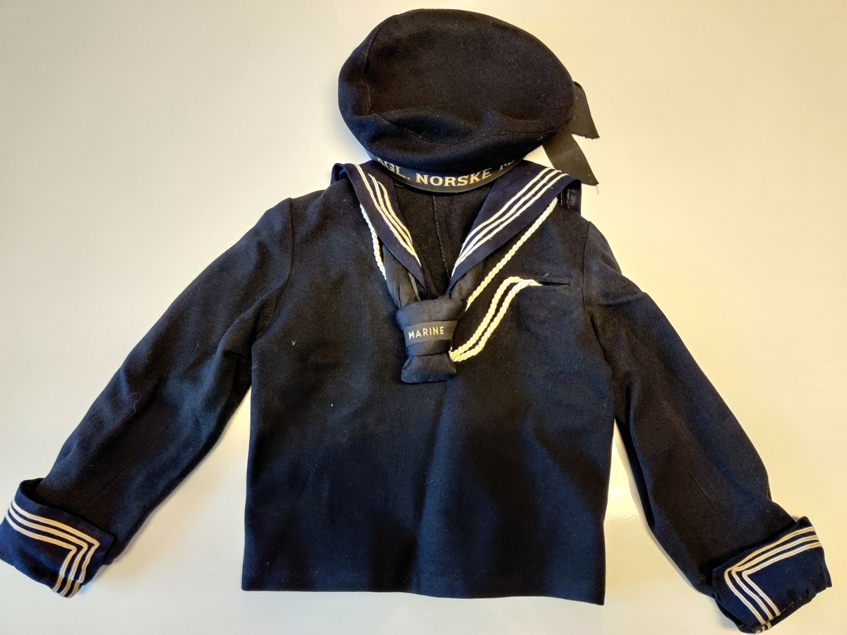 Sjømannsdress for barn. Består av trøye og hette. Farge: mørkeblå, kvitt rand.
Ullstoff, krage og slips bomullstoff.