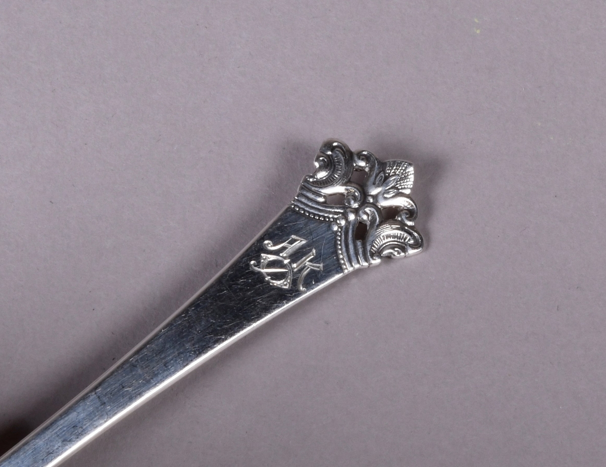 Skje av sølv med støpt dekor i enden av skaftet og ved overgang til skål. Mønsteret ved enden er gjennombrutt med fire rektangulære hull.