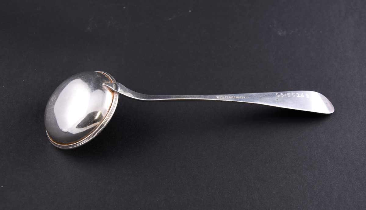 Serveringsskje av sølv. Skaftet er dekorert med innsenket mønster i form av et skjold med planteblader under og over.
