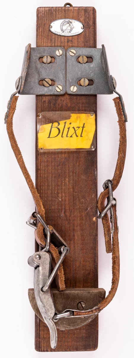 Skidbindningsprov. Metall på trä. Läderemmar. På ovansidan genomskinlig rektangulär plastskiva över gul etikett med text "Blixt". Elof Malmbergs firmamärke i metall.