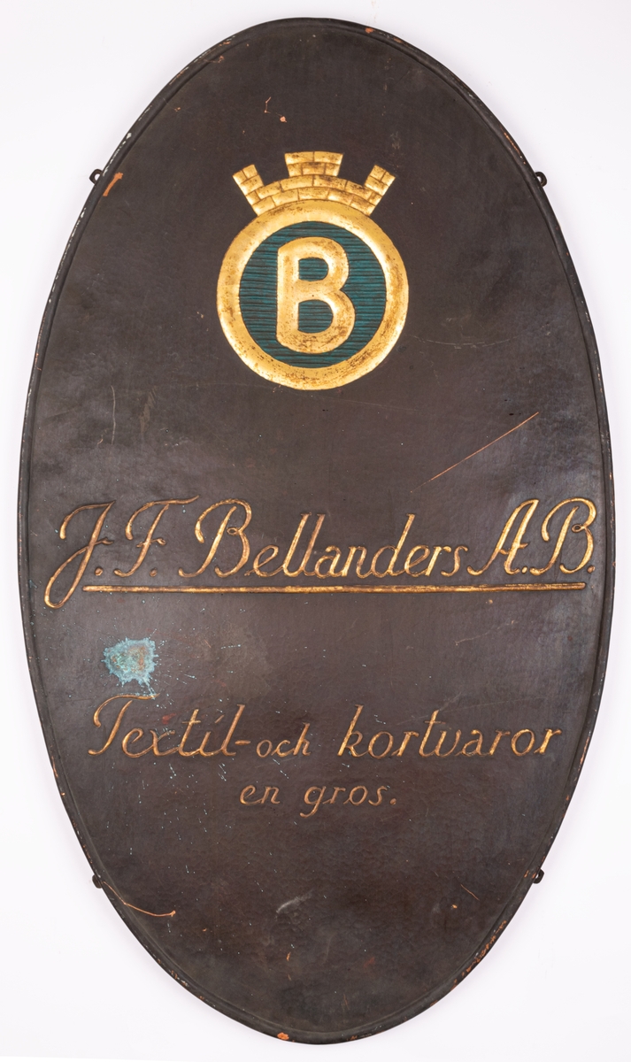 Skylt i tunn plåt. Ovalt stående format med bakåtböjd 2cm tjock kant. I plåten reliefpressade, förgyllda bokstäver mot mörkt grå fond. "J.F. Bellander A.B. textil och kortvaror. En gros.". Överst emblem med krönt förgylld ring, inneslutande förgyllt "B" mot blå fond. Fyra skruvfästen på skyltens sidor.