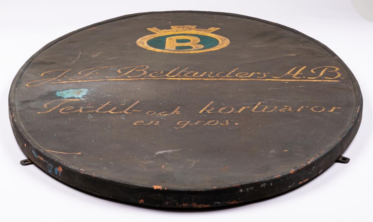 Skylt i tunn plåt. Ovalt stående format med bakåtböjd 2cm tjock kant. I plåten reliefpressade, förgyllda bokstäver mot mörkt grå fond. "J.F. Bellander A.B. textil och kortvaror. En gros.". Överst emblem med krönt förgylld ring, inneslutande förgyllt "B" mot blå fond. Fyra skruvfästen på skyltens sidor.