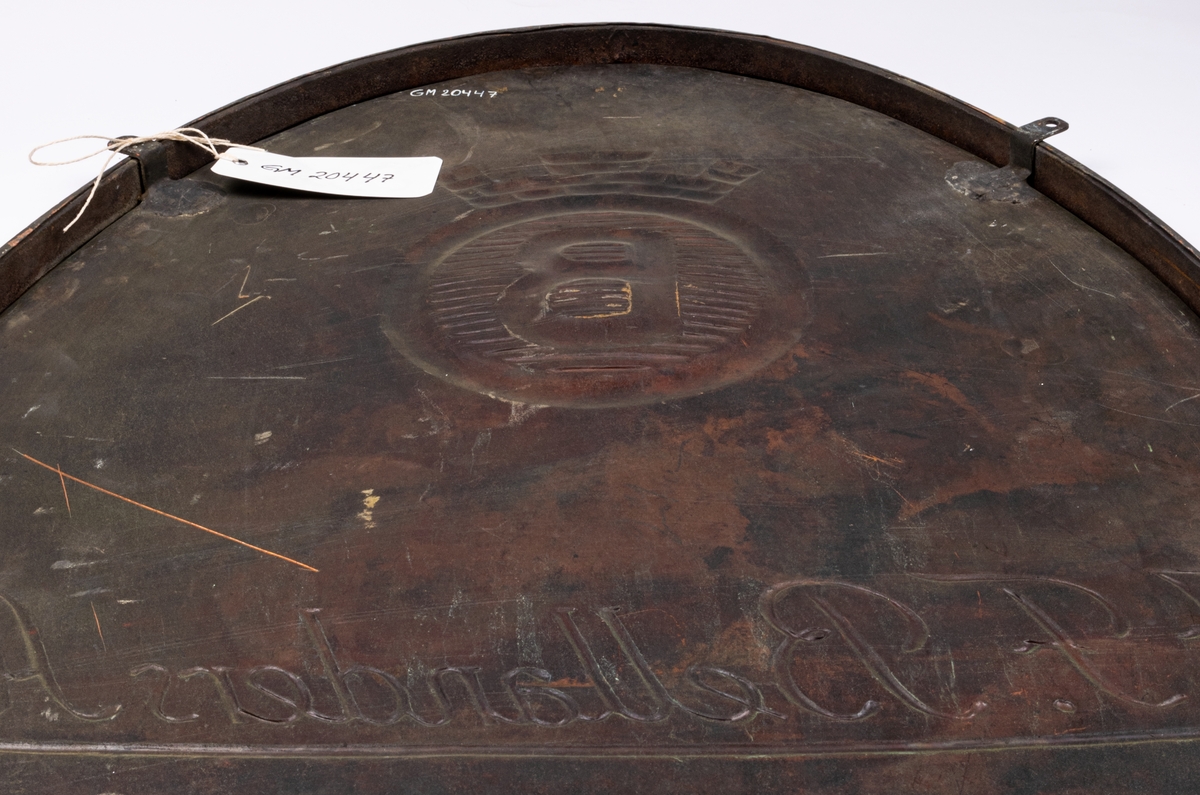 Skylt i tunn plåt. Ovalt stående format med bakåtböjd 2cm tjock kant. I plåten reliefpressade, förgyllda bokstäver mot mörkt grå fond. "J.F. Bellander A.B. textil och kortvaror. En gros.". Överst emblem med krönt förgylld ring, inneslutande förgyllt "B" mot blå fond. Fyra skruvfästen på skyltens sidor.