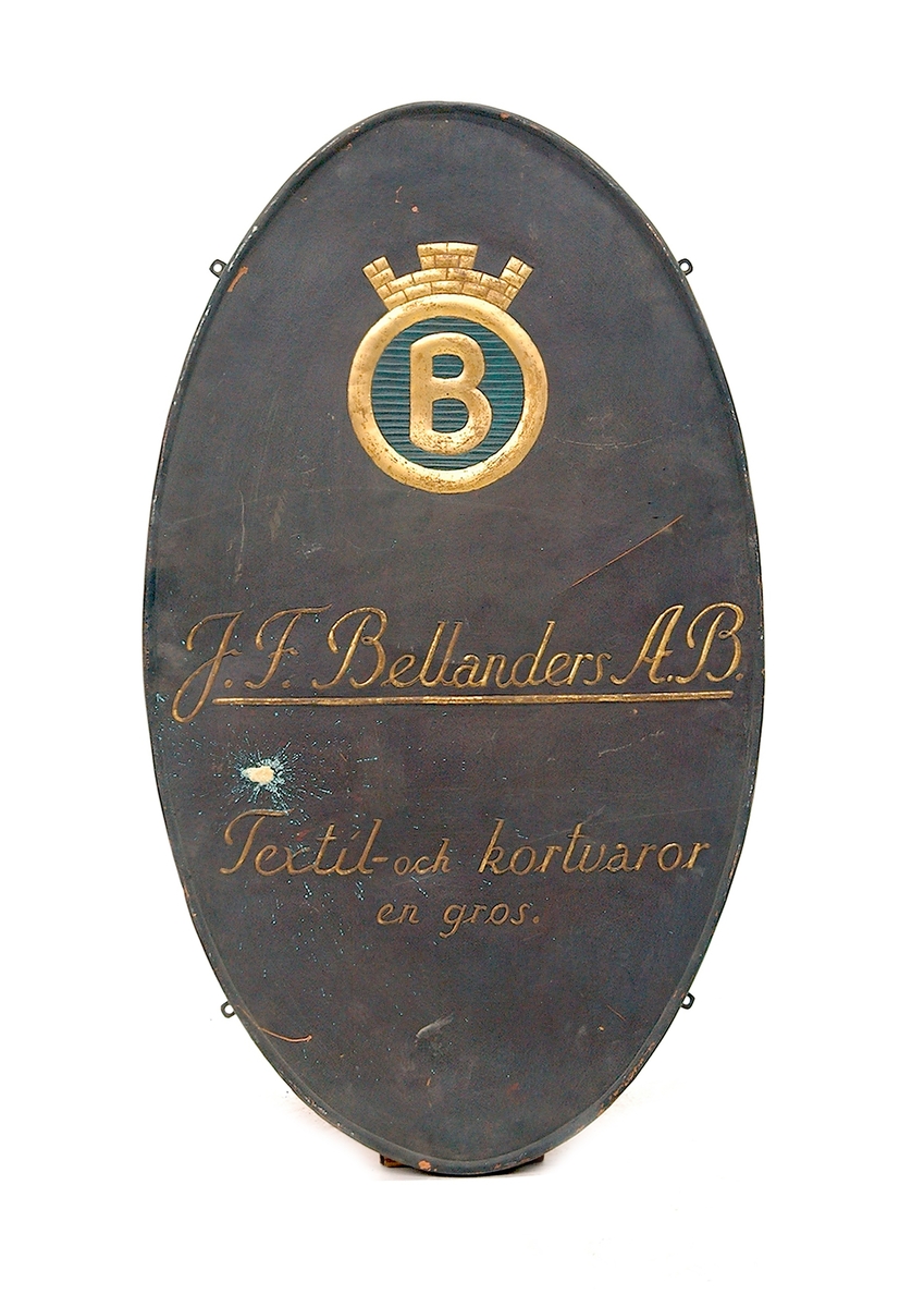 Skylt i tunn plåt. Ovalt stående format med bakåtböjd 2cm tjock kant. I plåten reliefpressade, förgyllda bokstäver mot mörkt grå fond. "J.F. Bellander A.B. textil och kortvaror. En gros.". Överst emblem med krönt förgylld ring, inneslutande förgyllt "B" mot blå fond. Fyra skruvfästen på skyltens sidor.