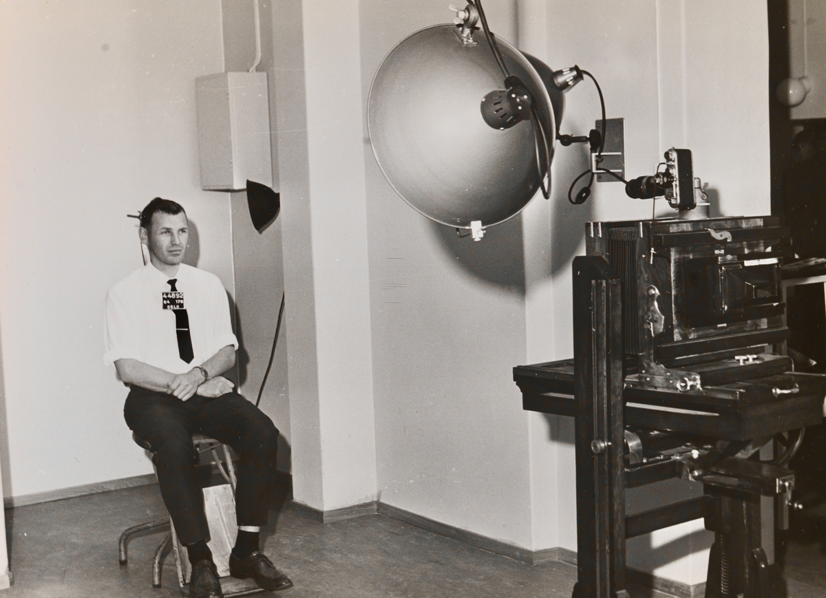 En mann demonstrerer metoden for forbryterfotografi. Han sitter på en spesialstol med nakkestøtte foran et stort fotoapparat.