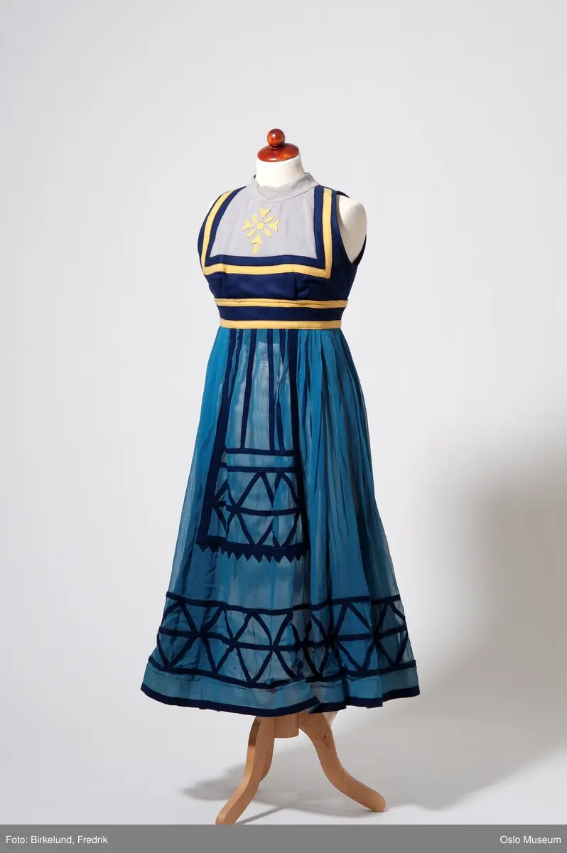 Ballettkjole: liv; mørkeblå, lysegrå og gult. Gule filtapplikasjoner. Ermeløs, høy hals. Skjørt; mellomblå chiffon med mørk blå filtapplikasjon. Lukkes med hekter og trykknapper i ryggen.