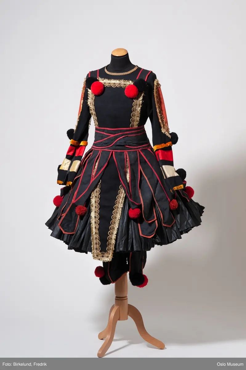 A: kort foldeskjørt med stor vidde. Sort med røde detaljer.
B: bukse, sort med røde detaljer. Stofftrukne knapper i siden ved kneet. Hvert kne er dekorert med en rød og en sort dusk. Lukkes med glidelås i front.
C: sort langermet genser. Innfelt oransje felt på hvert erme. Feltet er dekorert med gullbordog en rød og en sort dusk. Nedre del av begge ermer dekorstriper i rød, gul og gull samt en rødm en sort og en rødbrun dusk.
D: magebelte med "skjørt" Sort med røde detaljer. Nedre kant på "skjørt" er dekorert med annenhver rød og rødbrun dusk. Lukkes med tre hekter i rygg.
E: "vest/ kappe", trekkes over hodet med felt foran og bak. Kantet med gullbord. Hvert hjørne dekorert med en sort og en rød dusk.