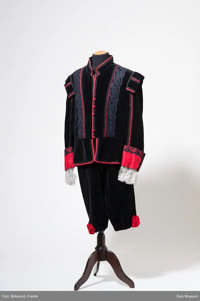 A: jakke i sort fløyel. Dekorert med sort blonder og pyntebånd samt røde pyntebånd. Røde stoftrukne knapper. Jakken lukkes med trykknapper i front. Hvite blonder langs håndledd. 
B: knebukser i sort fløyel. Lukkes med glidelås i siden. Dekorert med rød rosett nederst på hvert bukseben.
C: kappe i sort fløyel. Rødt for. Rød krave trukket med sorte blonder. Lukkes med sort bånd.