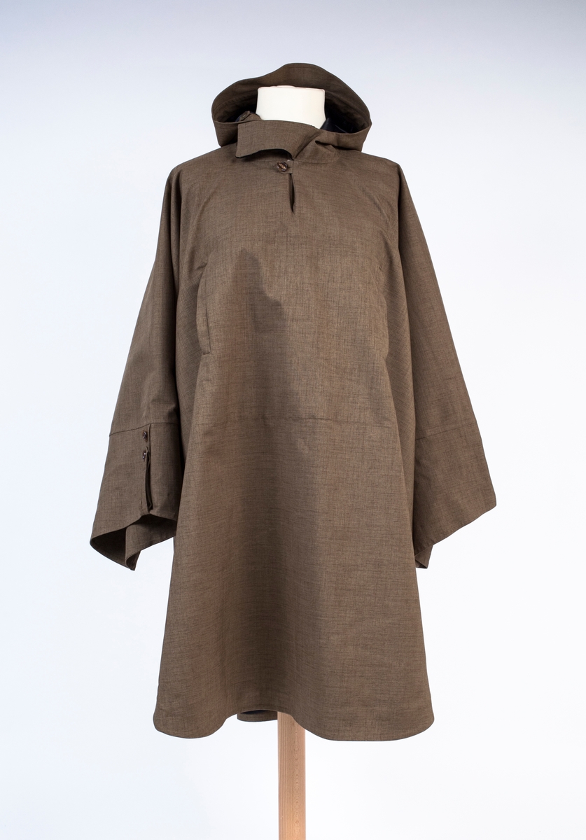 Unisex poncho med fast hette, løst belte og løst knyttebånd i korsryggen. Knelang foran, lengre bak. Primærsnitt med rett isatte ermer, i kombinasjon med "flaggermussnitt". Ingen skuldersømmer, kun sidesømmer og ermsømmer. Det ekstremt vide plagget, har kun korte, lett avsmalende ermer, vide ved håndleddet og lengst på undersiden. Oversiden av ermåpningen, er utstyrt med en dekorativ stropp og 2 knapper, der man kan regulere knappingen et hakk og få kortet ermet ørlite grann i overkant. To lavtstilte stikklommer i sidesømmen og to vertikaltstilte lommer på brystet. Brystlommene er ikke gjennomgående, men kan lukkes hver for seg med skjult glidelås. Plagget tres over hodet, og har en 11 cm dyp splitt i halsen, isatt en tre-delt kile eller motfold. Splitten er lukket med knapp og stoffhempe. I korsryggen kan midtre del av ryggstykket knyttes strammere ved hjelp av et smalt knyttebånd mellom to beltestropper. Påsydd treblads hette med skjerm og klaff i halsen. Hetten kan strammes omkring ansiktet ved hjelp av et smalt bånd gjennom løpegang i fronten. Dette er kneppet fast med trykknapp i hver ende, med et ekstra par trykknapper lenger ned, for en strammere posisjon. Må da eventuelt knytes etter stramming. Innsiden av halsklaffen er fôret med stoff av kashmirull, til beskyttelse mot kulde i halsen. Klaffen har trinnløs regulering ved hjelp av borrelås. Hele plagget er helfôret med blankt syntetstoff. På varefaktalappen i høyre sidesøm, er påsydd 2 reserveknapper. På innsiden av forstykket, i låshøyde, er festet to brede, horisontale løkker av elastikk, antagelig for å tre bena gjennom og holde frontflaket på plass ved vind eller sykling. Langt belte følger med. Dette er sydd med skråstilt spiss i ene enden og to stropper for itreing i motsatt side. Ingen beltespenne.