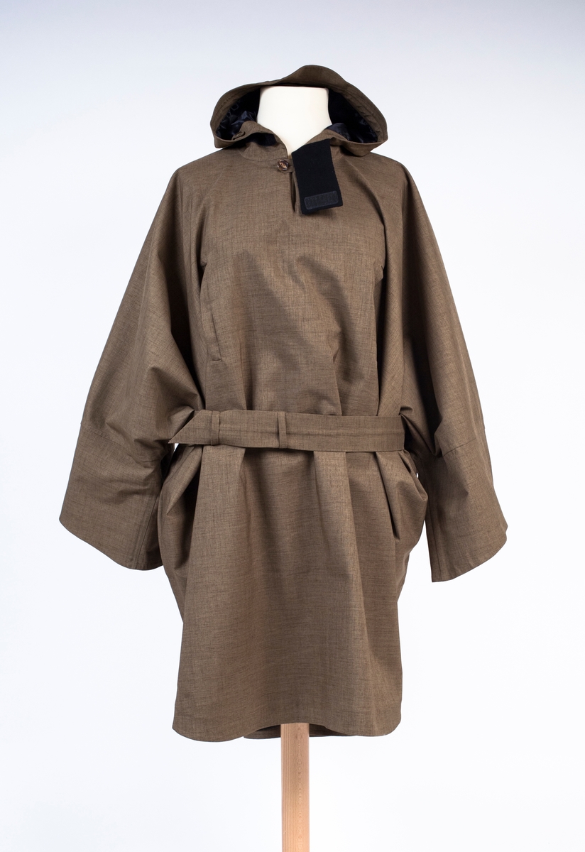 Unisex poncho med fast hette, løst belte og løst knyttebånd i korsryggen. Knelang foran, lengre bak. Primærsnitt med rett isatte ermer, i kombinasjon med "flaggermussnitt". Ingen skuldersømmer, kun sidesømmer og ermsømmer. Det ekstremt vide plagget, har kun korte, lett avsmalende ermer, vide ved håndleddet og lengst på undersiden. Oversiden av ermåpningen, er utstyrt med en dekorativ stropp og 2 knapper, der man kan regulere knappingen et hakk og få kortet ermet ørlite grann i overkant. To lavtstilte stikklommer i sidesømmen og to vertikaltstilte lommer på brystet. Brystlommene er ikke gjennomgående, men kan lukkes hver for seg med skjult glidelås. Plagget tres over hodet, og har en 11 cm dyp splitt i halsen, isatt en tre-delt kile eller motfold. Splitten er lukket med knapp og stoffhempe. I korsryggen kan midtre del av ryggstykket knyttes strammere ved hjelp av et smalt knyttebånd mellom to beltestropper. Påsydd treblads hette med skjerm og klaff i halsen. Hetten kan strammes omkring ansiktet ved hjelp av et smalt bånd gjennom løpegang i fronten. Dette er kneppet fast med trykknapp i hver ende, med et ekstra par trykknapper lenger ned, for en strammere posisjon. Må da eventuelt knytes etter stramming. Innsiden av halsklaffen er fôret med stoff av kashmirull, til beskyttelse mot kulde i halsen. Klaffen har trinnløs regulering ved hjelp av borrelås. Hele plagget er helfôret med blankt syntetstoff. På varefaktalappen i høyre sidesøm, er påsydd 2 reserveknapper. På innsiden av forstykket, i låshøyde, er festet to brede, horisontale løkker av elastikk, antagelig for å tre bena gjennom og holde frontflaket på plass ved vind eller sykling. Langt belte følger med. Dette er sydd med skråstilt spiss i ene enden og to stropper for itreing i motsatt side. Ingen beltespenne.