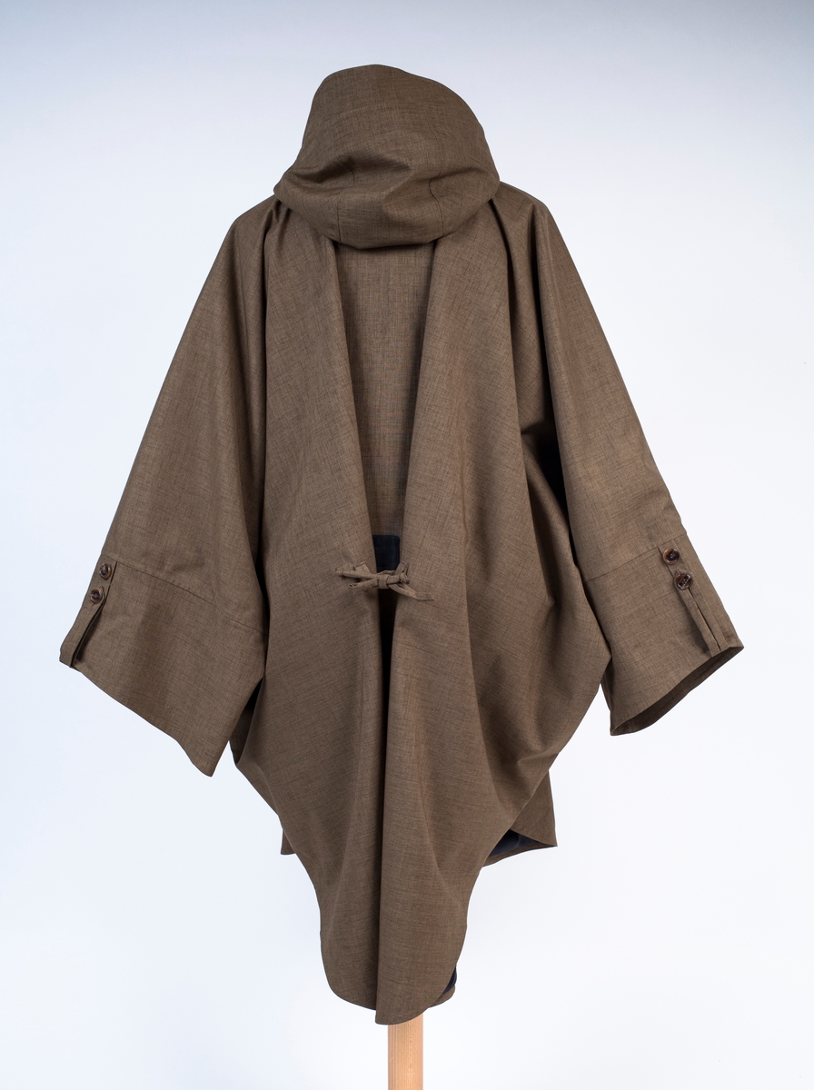 Unisex poncho med fast hette, løst belte og løst knyttebånd i korsryggen. Knelang foran, lengre bak. Primærsnitt med rett isatte ermer, i kombinasjon med "flaggermussnitt". Ingen skuldersømmer, kun sidesømmer og ermsømmer. Det ekstremt vide plagget, har kun korte, lett avsmalende ermer, vide ved håndleddet og lengst på undersiden. Oversiden av ermåpningen, er utstyrt med en dekorativ stropp og 2 knapper, der man kan regulere knappingen et hakk og få kortet ermet ørlite grann i overkant. To lavtstilte stikklommer i sidesømmen og to vertikaltstilte lommer på brystet. Brystlommene er ikke gjennomgående, men kan lukkes hver for seg med skjult glidelås. Plagget tres over hodet, og har en 11 cm dyp splitt i halsen, isatt en tre-delt kile eller motfold. Splitten er lukket med knapp og stoffhempe. I korsryggen kan midtre del av ryggstykket knyttes strammere ved hjelp av et smalt knyttebånd mellom to beltestropper. Påsydd treblads hette med skjerm og klaff i halsen. Hetten kan strammes omkring ansiktet ved hjelp av et smalt bånd gjennom løpegang i fronten. Dette er kneppet fast med trykknapp i hver ende, med et ekstra par trykknapper lenger ned, for en strammere posisjon. Må da eventuelt knytes etter stramming. Innsiden av halsklaffen er fôret med stoff av kashmirull, til beskyttelse mot kulde i halsen. Klaffen har trinnløs regulering ved hjelp av borrelås. Hele plagget er helfôret med blankt syntetstoff. På varefaktalappen i høyre sidesøm, er påsydd 2 reserveknapper. På innsiden av forstykket, i låshøyde, er festet to brede, horisontale løkker av elastikk, antagelig for å tre bena gjennom og holde frontflaket på plass ved vind eller sykling. Langt belte følger med. Dette er sydd med skråstilt spiss i ene enden og to stropper for itreing i motsatt side. Ingen beltespenne.