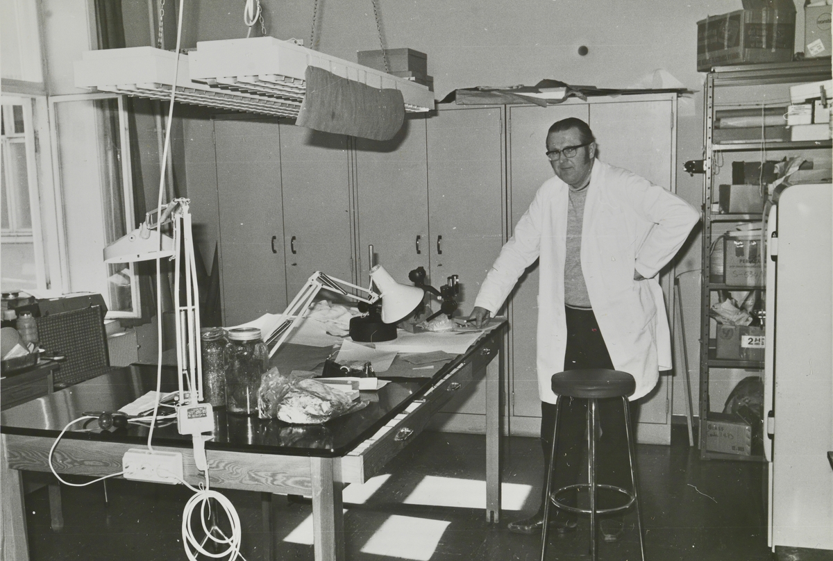 En mann i labfrakk står ved et arbeidsbord.