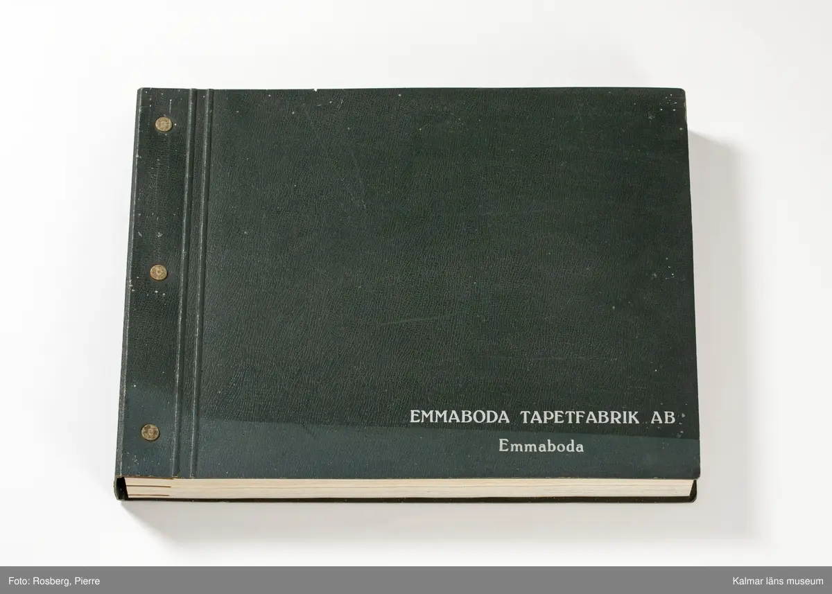 KLM 46599:3. Tapetkatalog. Katalog med pärm av grön papp eller kartong. Bunden med hjälp av tre metallnitar. Text i silver på pärmens framsida: EMMABODA TAPETFABRIK AB Emmaboda. Katalogen innehåller en mängd olika plastförstärkta tapetprover tillverkade av papper, med olika strukturer, färger och mönster, moderna under 1950 och 1960-talen.