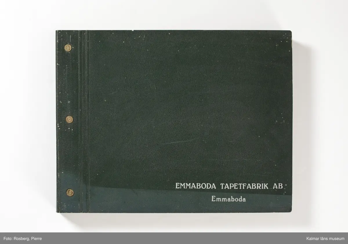 KLM 46599:3. Tapetkatalog. Katalog med pärm av grön papp eller kartong. Bunden med hjälp av tre metallnitar. Text i silver på pärmens framsida: EMMABODA TAPETFABRIK AB Emmaboda. Katalogen innehåller en mängd olika plastförstärkta tapetprover tillverkade av papper, med olika strukturer, färger och mönster, moderna under 1950 och 1960-talen.