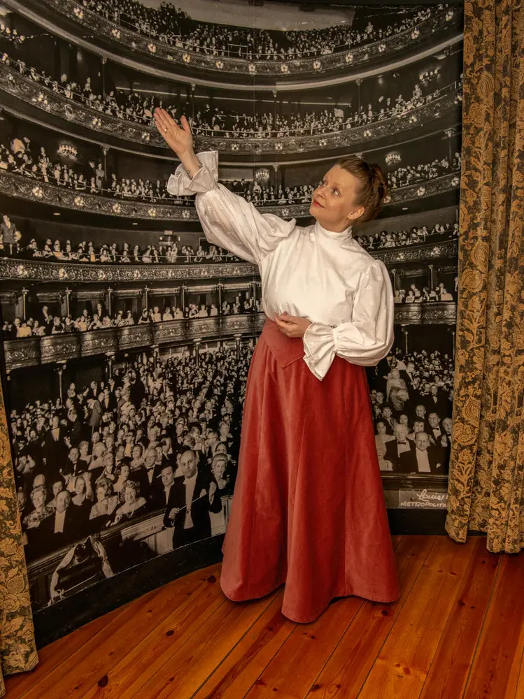 Ung kvinne med hårtopp og historisk drakt bestående av hvit bluse og rødt, fotsidt skjørt står foran et stort veggbilde fra The Metropolitan og guider om Kirsten Flagstads gjennombrudd.