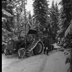 En skogstraktor står i en skogsglänta. Den har terränghjul. Det är snö på marken. Två män lastar trädstammar på en kärra.