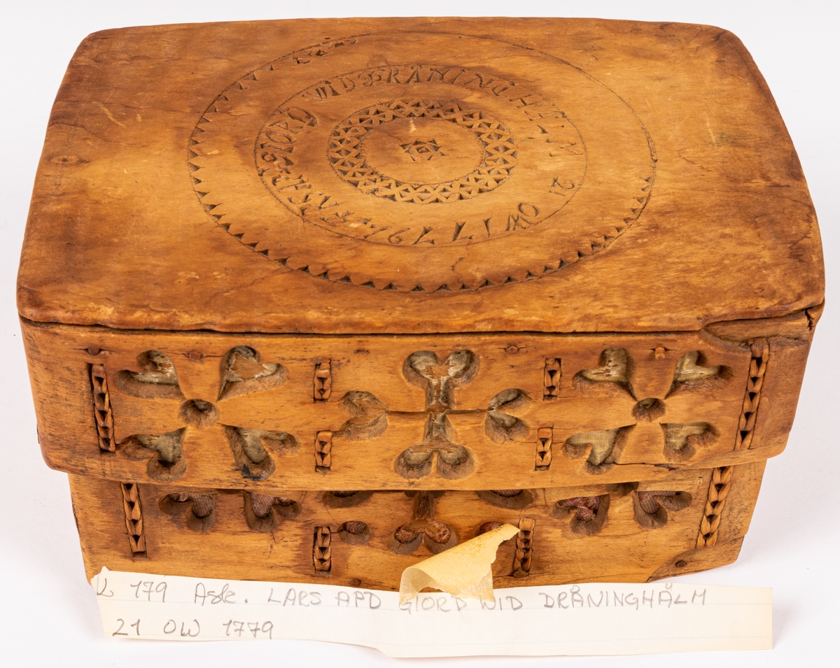 Ask med trycklock, fyrsidig, av trä, utskuret på en sida samt på locket: "LARS PDS GJORD WID DRÅNINGHÅLM 21 OW 1779". Svepteknik. På ena sidan av lådan och på dess lock är övre skiktet av svepet genombrutet av utskurna fyrarmade figurer med rött resp. vitt papper under.