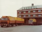 Lager, skeppningskaj. Två scania lastbilar, den ena orange och med släp, den andra grön med gult kapell.  lastar/lossar vid kajen lagret. Röd tegelbyggnad med LÄKEROL skylt på taket.