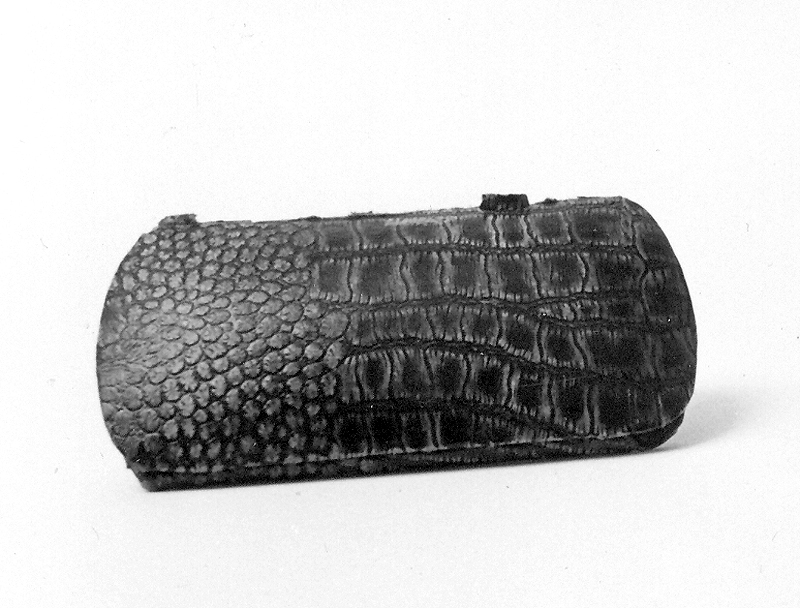 Glasögonfodral av plåt, tyg och papp. Plåtfodral utvändigt. Klätt med papp i brun läderimitation. Uppfällbart lock. Rektangulär form. Invändigt klätt med grönt tyg. Metalletikett i guldfärg på insidan av locket. Märkt: "GUST NORMAN, UR & OPTIK, VARBERG, TEL. 222". Läderimitationen trasig vid gångjärnen till locket.