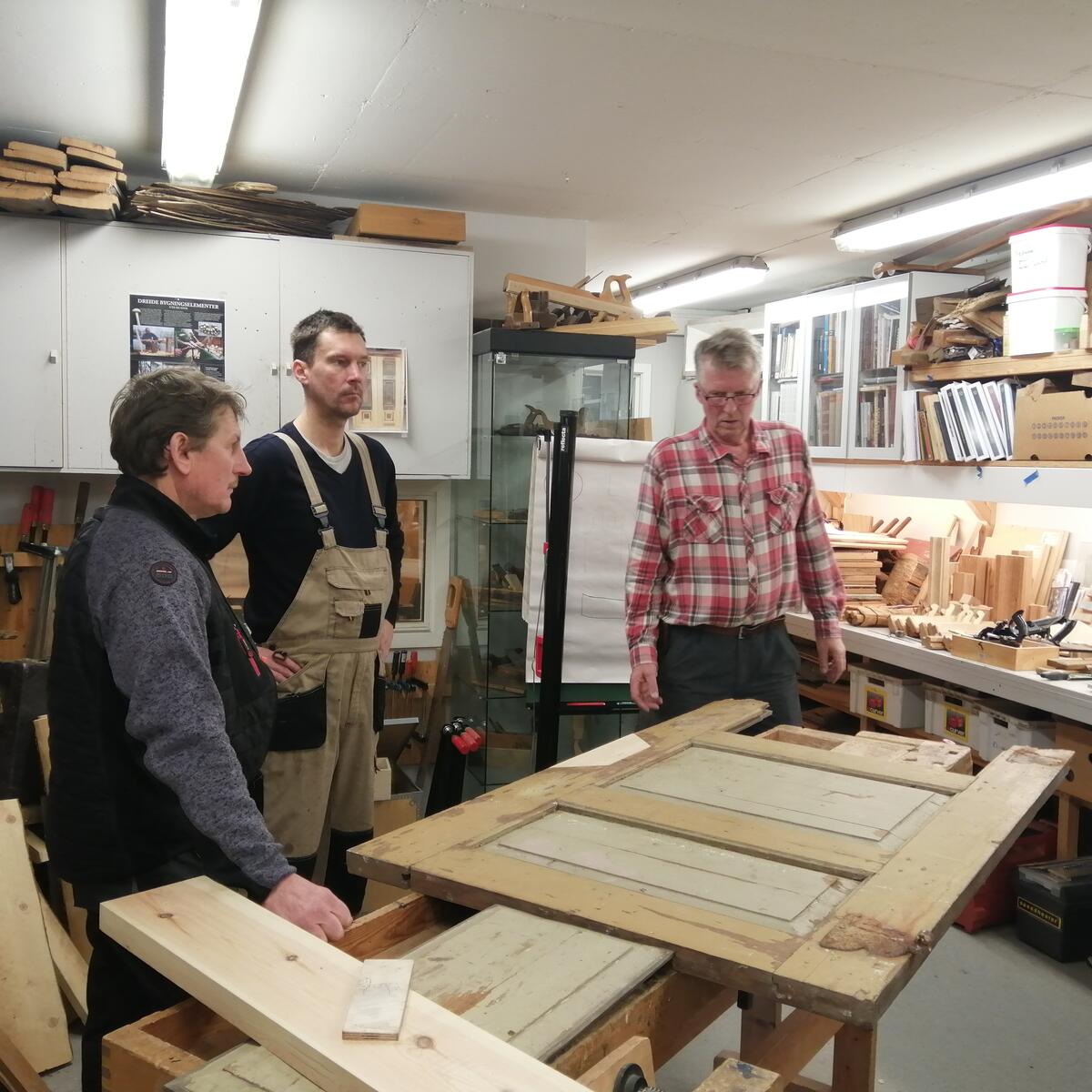 Tre håndverkere i et verksted står samlet rundt en gammel dør.