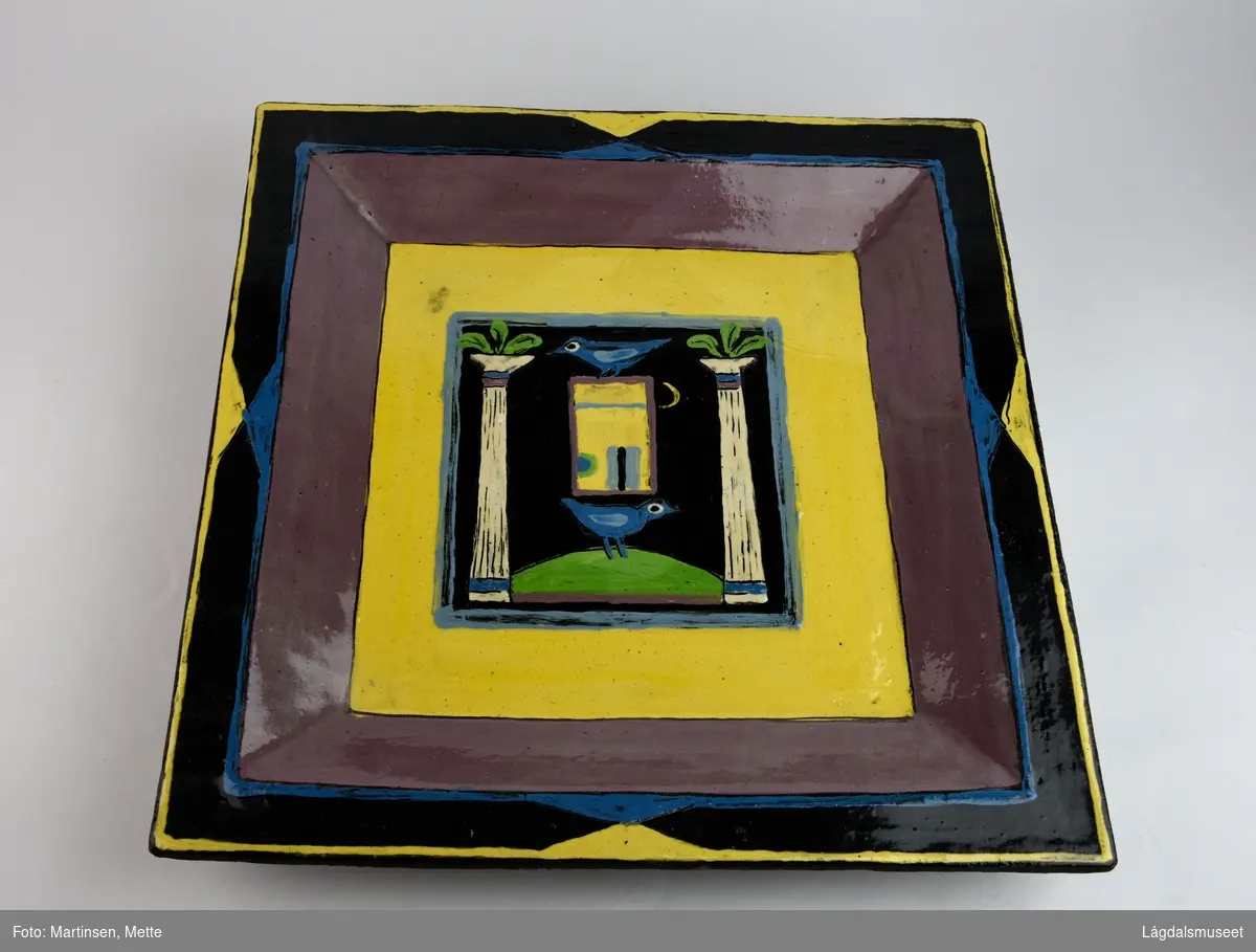 Veggfat i keramikk. Kvadratisk ramme i fatets speil med figurativt motiv. To blå fugler står henholdsvis over og under en dør eller vindu. På hver side søyler med tre palmeblader på toppen. Motivet er rammet inn av tre bredere rammer i gult, brunt og svart.