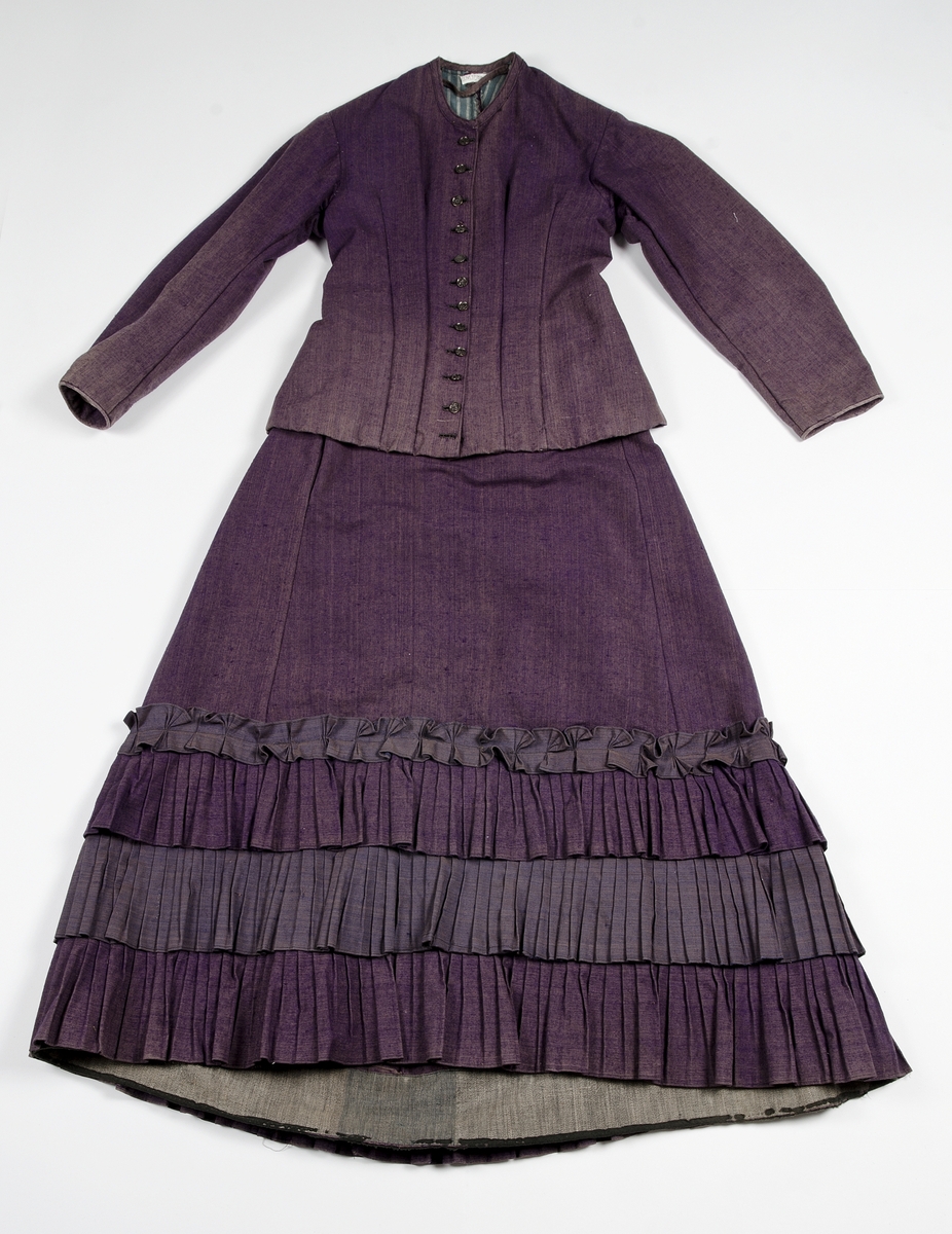 Av tuskaftat lila och brunt ylletyg med volanger av blåbrunt tyg. Jackan knäppt med tolv svartlackerade metallknappar med blommotiv. Fodrad med blågrön och gulrandigt bomullstyg. Ärmarna med vit linnelärft med tryckt mönster av röda blommor. Kjolens nederkant skodd med grönt bomullsband.