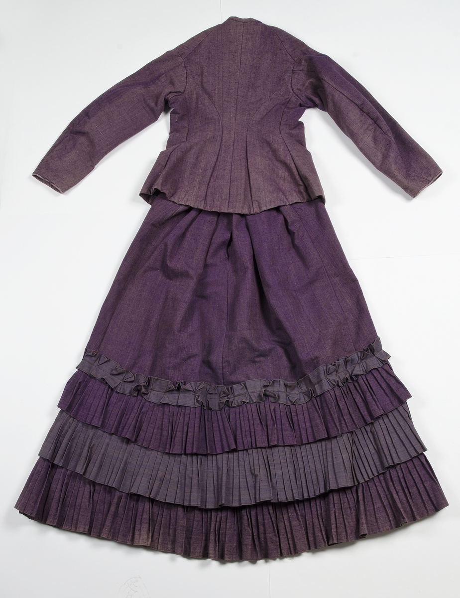 Av tuskaftat lila och brunt ylletyg med volanger av blåbrunt tyg. Jackan knäppt med tolv svartlackerade metallknappar med blommotiv. Fodrad med blågrön och gulrandigt bomullstyg. Ärmarna med vit linnelärft med tryckt mönster av röda blommor. Kjolens nederkant skodd med grönt bomullsband.