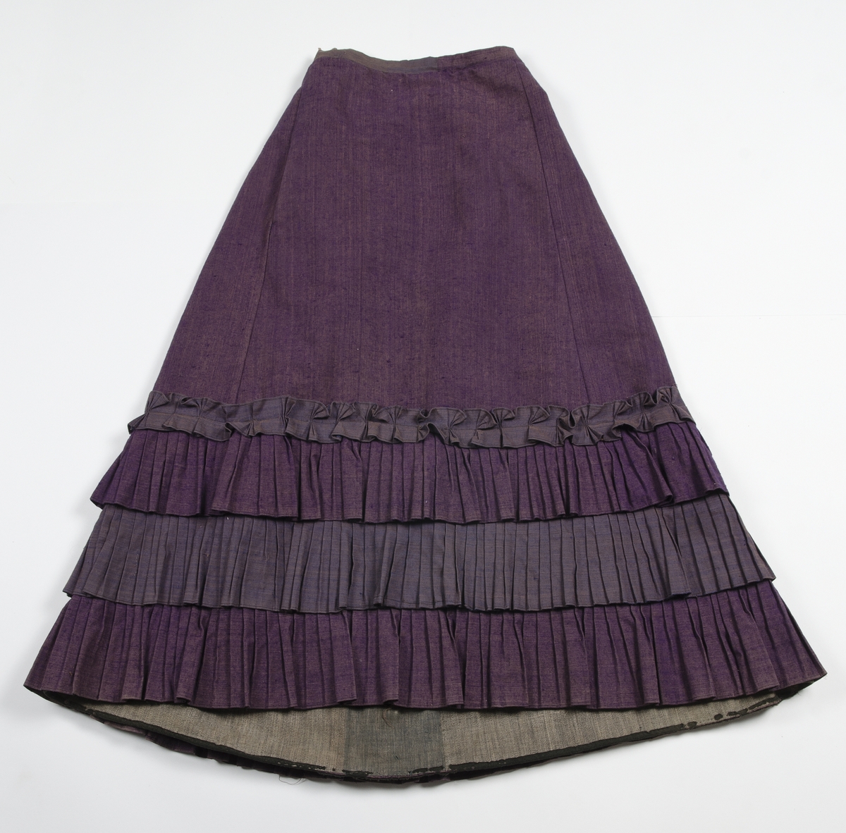 Av tuskaftat lila och brunt ylletyg med volanger av blåbrunt tyg. Jackan knäppt med tolv svartlackerade metallknappar med blommotiv. Fodrad med blågrön och gulrandigt bomullstyg. Ärmarna med vit linnelärft med tryckt mönster av röda blommor. Kjolens nederkant skodd med grönt bomullsband.