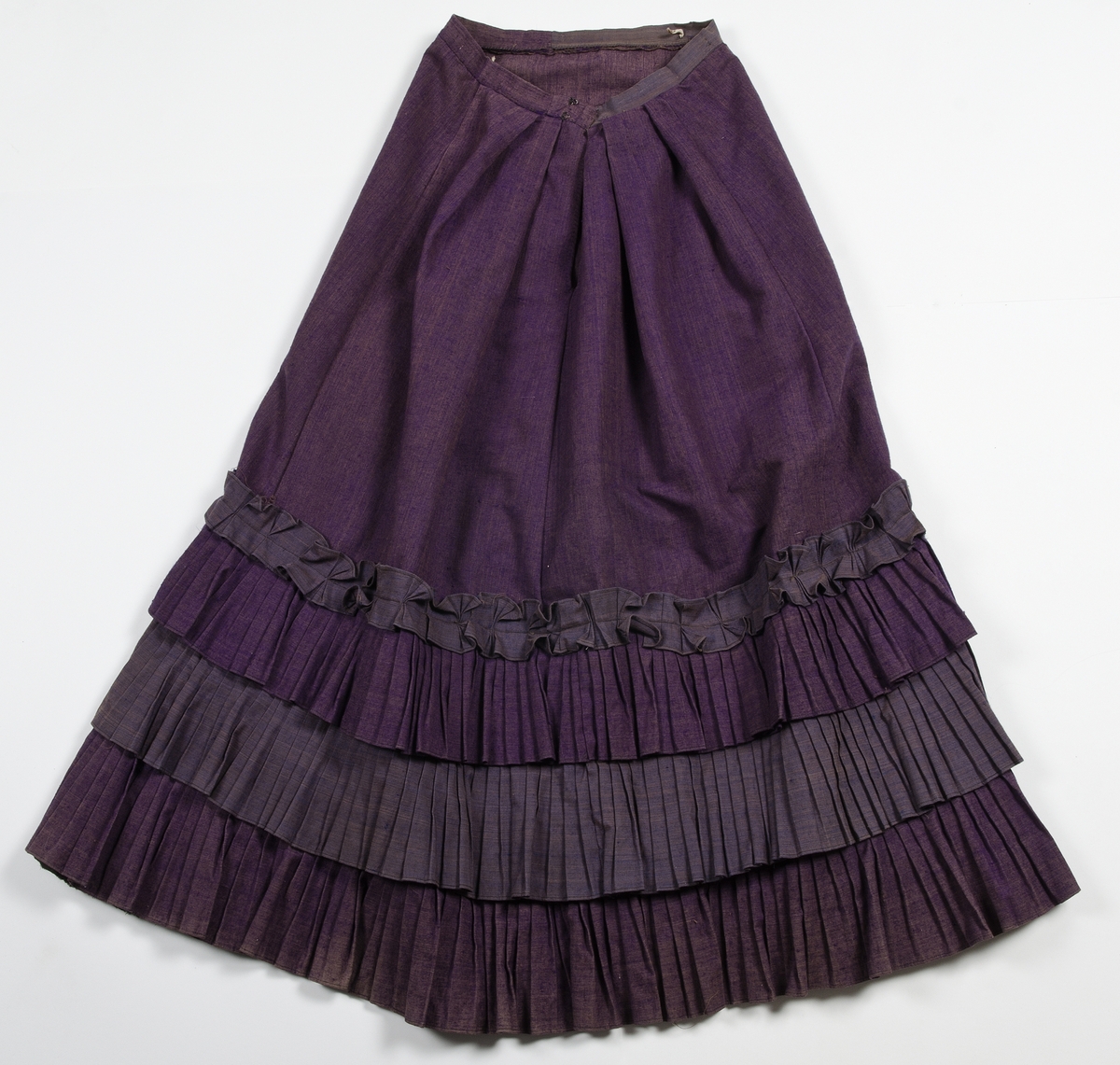 Av tuskaftat lila och brunt ylletyg med volanger av blåbrunt tyg. Jackan knäppt med tolv svartlackerade metallknappar med blommotiv. Fodrad med blågrön och gulrandigt bomullstyg. Ärmarna med vit linnelärft med tryckt mönster av röda blommor. Kjolens nederkant skodd med grönt bomullsband.