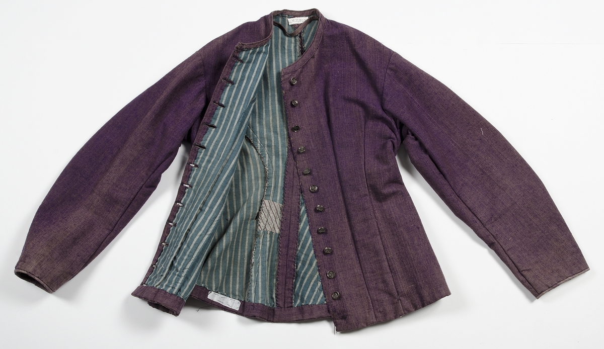 Av tuskaftat lila och brunt ylletyg med volanger av blåbrunt tyg. Jackan knäppt med tolv svartlackerade metallknappar med blommotiv. Fodrad med blågrön och gulrandigt bomullstyg. Ärmarna med vit linnelärft med tryckt mönster av röda blommor. Kjolens nederkant skodd med grönt bomullsband.