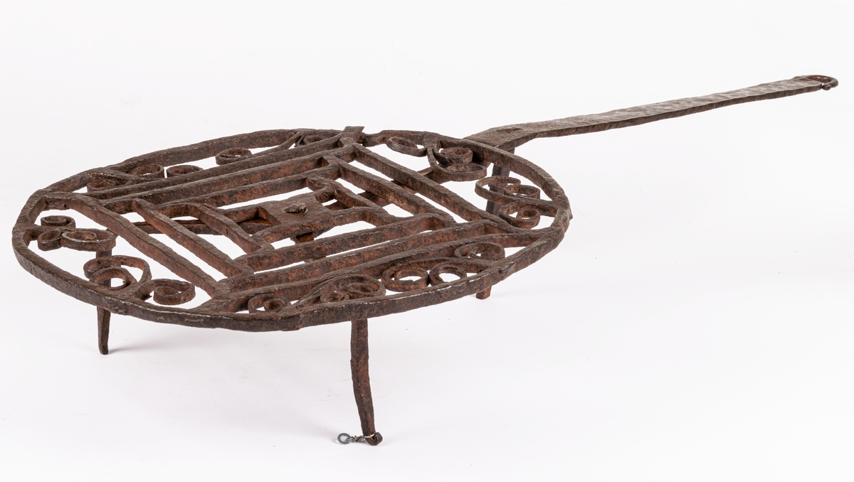 Halster av järn. Rund platta av hopsmidda tenar, jämte furkanter omgivna av cirkel och bladformiga ornament. Långt skaft, tre fötter.
