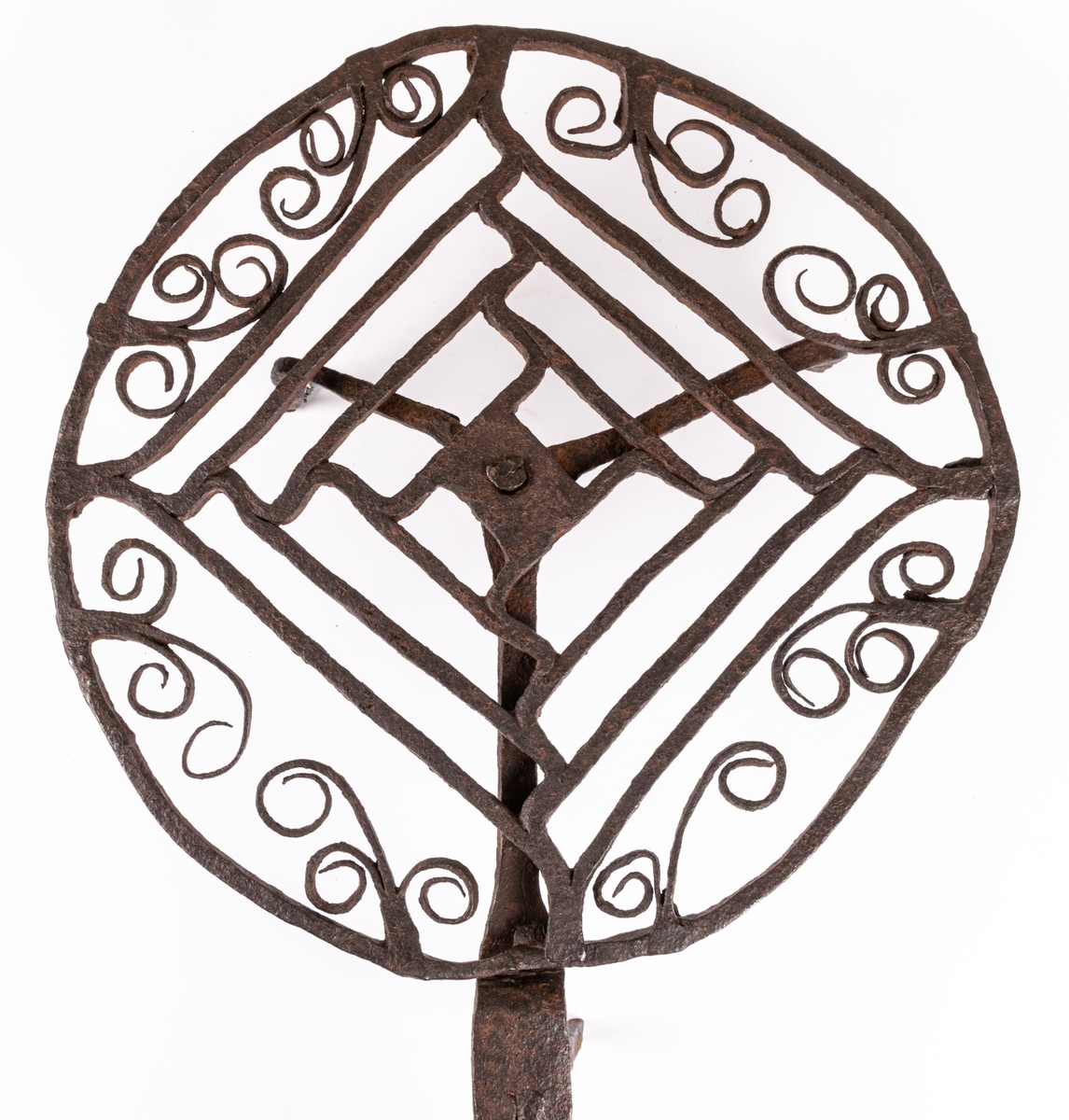 Halster av järn. Rund platta av hopsmidda tenar, jämte furkanter omgivna av cirkel och bladformiga ornament. Långt skaft, tre fötter.