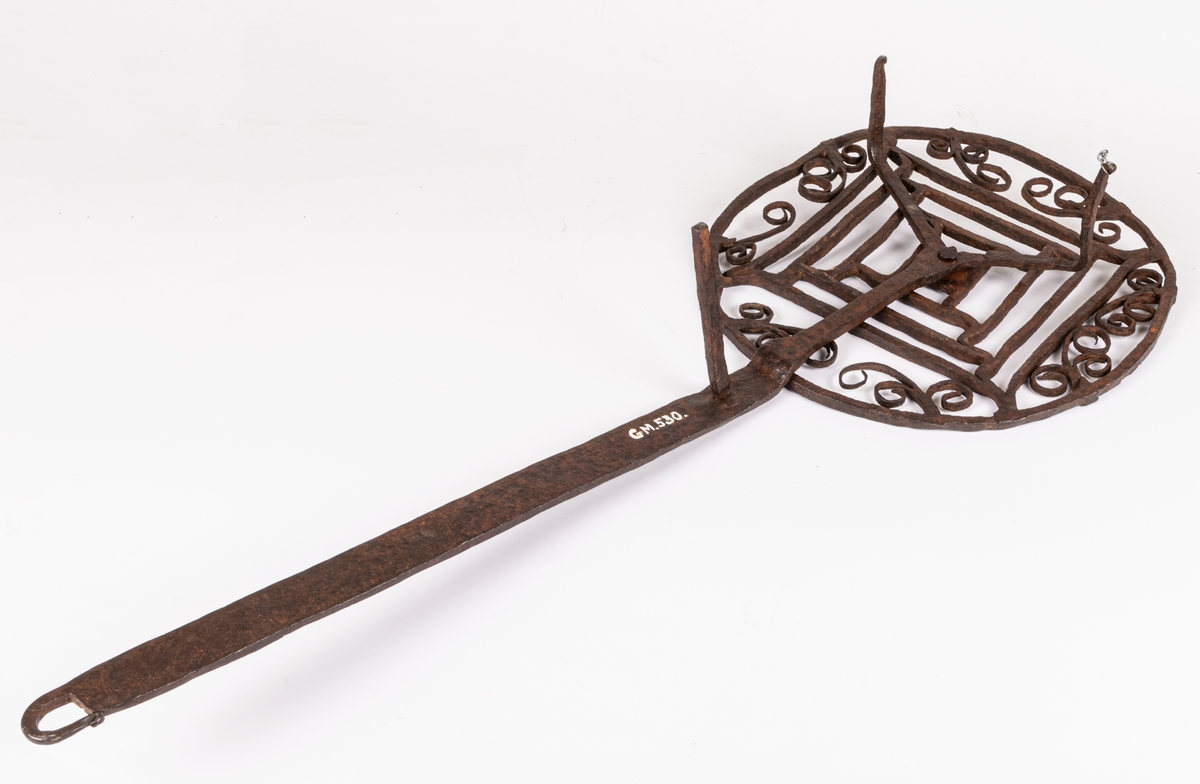 Halster av järn. Rund platta av hopsmidda tenar, jämte furkanter omgivna av cirkel och bladformiga ornament. Långt skaft, tre fötter.
