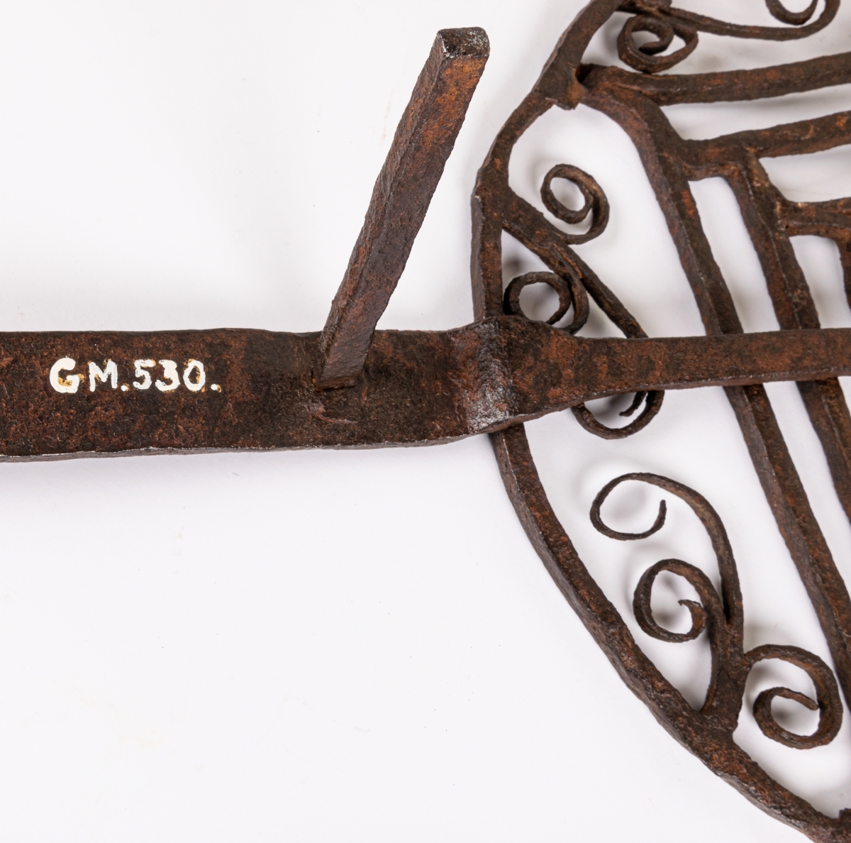 Halster av järn. Rund platta av hopsmidda tenar, jämte furkanter omgivna av cirkel och bladformiga ornament. Långt skaft, tre fötter.