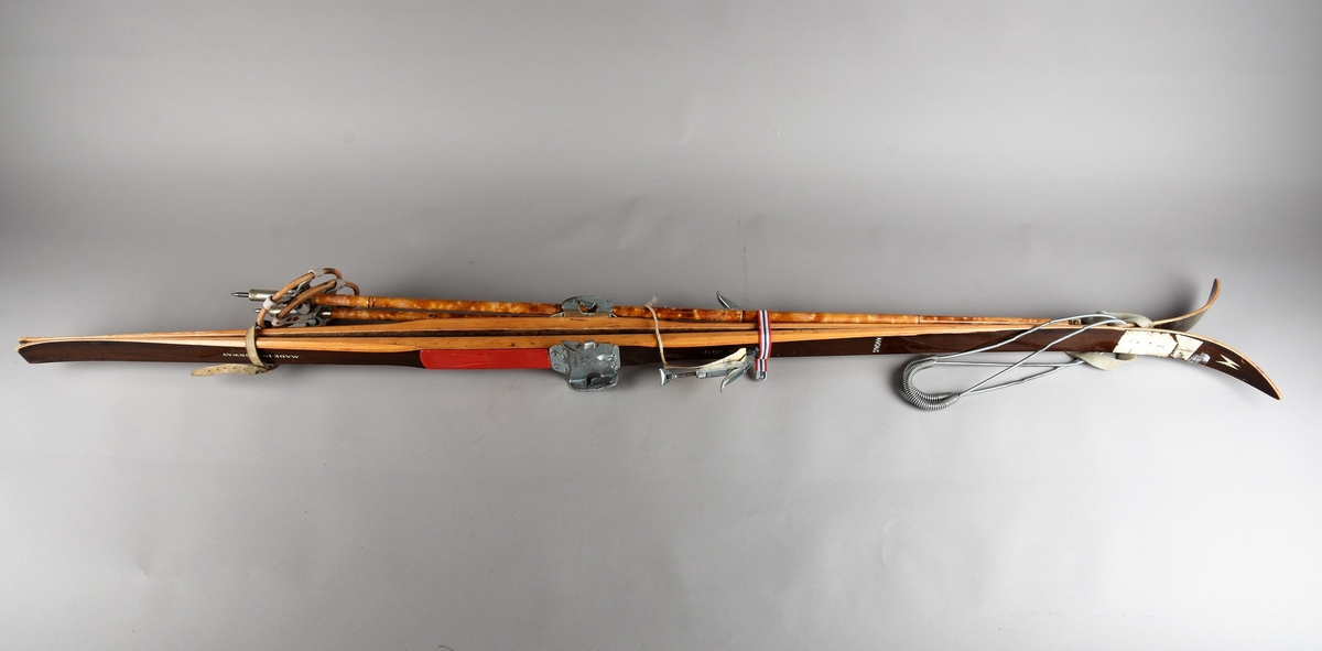 Eit par ski av tre. Kandaharbinding. Eit par skistavar. To reimer til å halde det saman. Ein reisegodslapp heng fast i B.