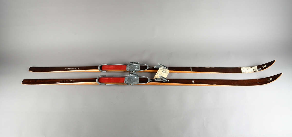 Eit par ski av tre. Kandaharbinding. Eit par skistavar. To reimer til å halde det saman. Ein reisegodslapp heng fast i B.