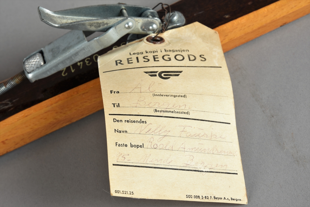 Eit par ski av tre. Kandaharbinding. Eit par skistavar. To reimer til å halde det saman. Ein reisegodslapp heng fast i B.