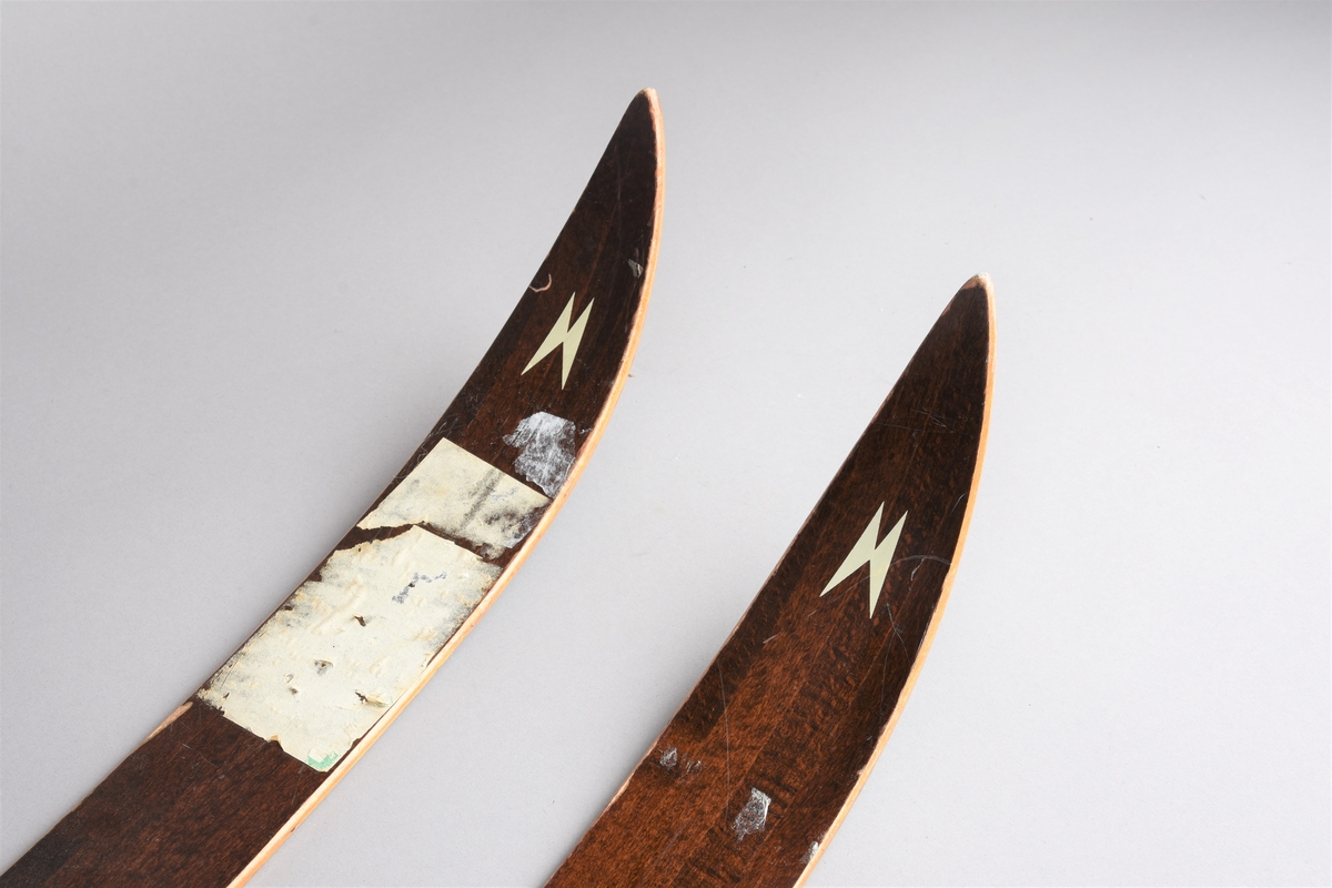 Eit par ski av tre. Kandaharbinding. Eit par skistavar. To reimer til å halde det saman. Ein reisegodslapp heng fast i B.