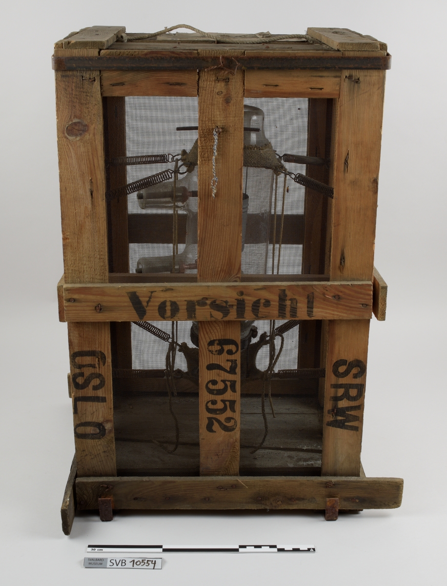 Kasse av tre. Bunnen og toppen er heldekkene, mens sidene er laget av langsgående trebord med mellomrom, avstivet med kortere bord på tvers i bunn og på midten. På innsiden av trebordene er det montert netting. Inni kassen er det montert et kortbølgeradiorør av glass og metall. Røret er suspendert i midten av kassen ved hjelp av to tekstilbiter, én i hver ende. Disse er holdt på plass av åtte metallfjærer, fire i hver ende, som er montert i kassens hjørner. Fjærene er montert ca. midt på kassens øverst halvdel og midt på kassens nederste halvdel. På den ene siden av kassens bunn er det montert to 90-graders metallbraketter. Langs toppen av den ene siden av kassen er det montert en lengre metallbrakett. I toppen av kassen er det festet tau og en papirlapp. På kassens utside er det trykket svart tekst. Trebordene holdes sammen med spiker og stifter av metall.