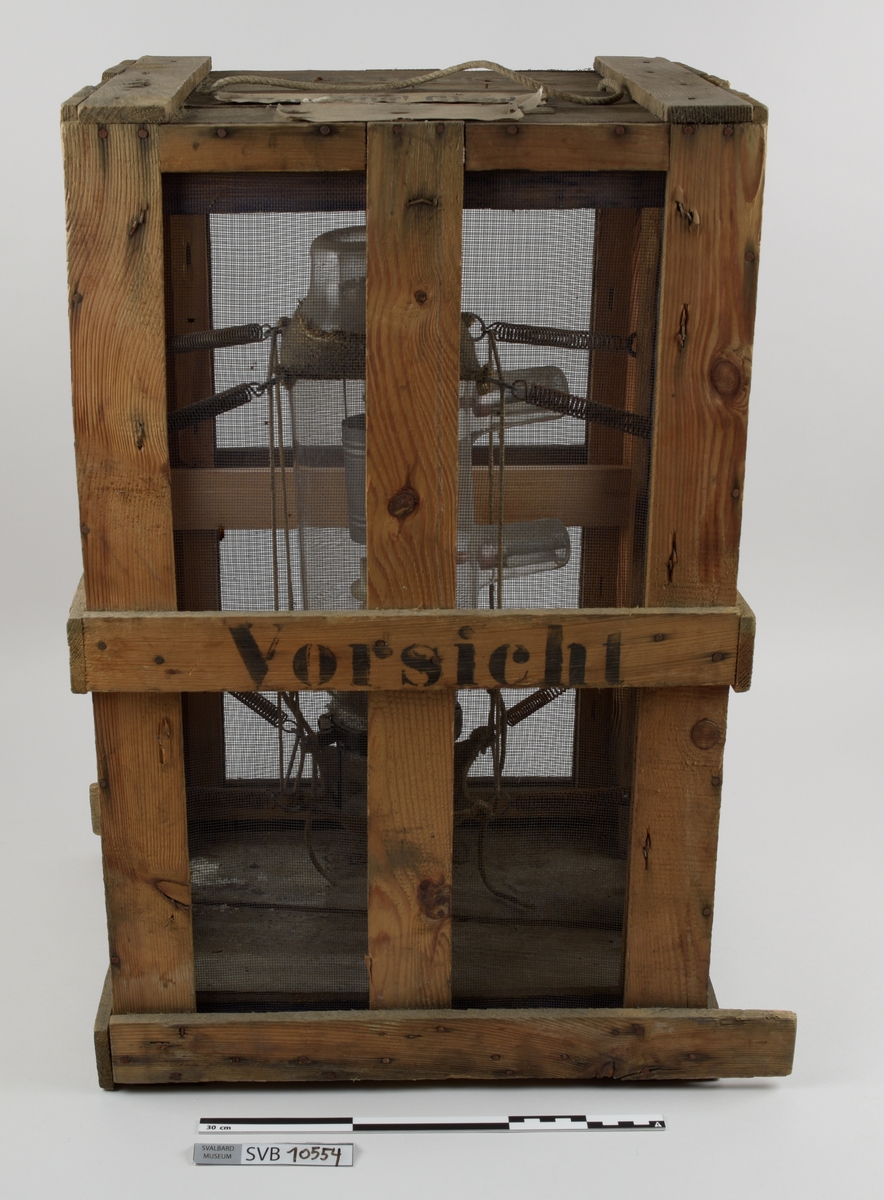 Kasse av tre. Bunnen og toppen er heldekkene, mens sidene er laget av langsgående trebord med mellomrom, avstivet med kortere bord på tvers i bunn og på midten. På innsiden av trebordene er det montert netting. Inni kassen er det montert et kortbølgeradiorør av glass og metall. Røret er suspendert i midten av kassen ved hjelp av to tekstilbiter, én i hver ende. Disse er holdt på plass av åtte metallfjærer, fire i hver ende, som er montert i kassens hjørner. Fjærene er montert ca. midt på kassens øverst halvdel og midt på kassens nederste halvdel. På den ene siden av kassens bunn er det montert to 90-graders metallbraketter. Langs toppen av den ene siden av kassen er det montert en lengre metallbrakett. I toppen av kassen er det festet tau og en papirlapp. På kassens utside er det trykket svart tekst. Trebordene holdes sammen med spiker og stifter av metall.