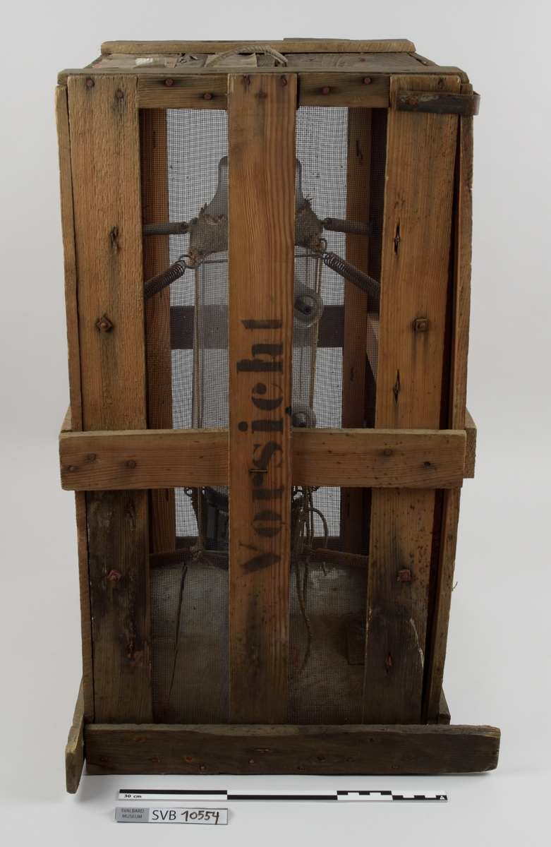 Kasse av tre. Bunnen og toppen er heldekkene, mens sidene er laget av langsgående trebord med mellomrom, avstivet med kortere bord på tvers i bunn og på midten. På innsiden av trebordene er det montert netting. Inni kassen er det montert et kortbølgeradiorør av glass og metall. Røret er suspendert i midten av kassen ved hjelp av to tekstilbiter, én i hver ende. Disse er holdt på plass av åtte metallfjærer, fire i hver ende, som er montert i kassens hjørner. Fjærene er montert ca. midt på kassens øverst halvdel og midt på kassens nederste halvdel. På den ene siden av kassens bunn er det montert to 90-graders metallbraketter. Langs toppen av den ene siden av kassen er det montert en lengre metallbrakett. I toppen av kassen er det festet tau og en papirlapp. På kassens utside er det trykket svart tekst. Trebordene holdes sammen med spiker og stifter av metall.